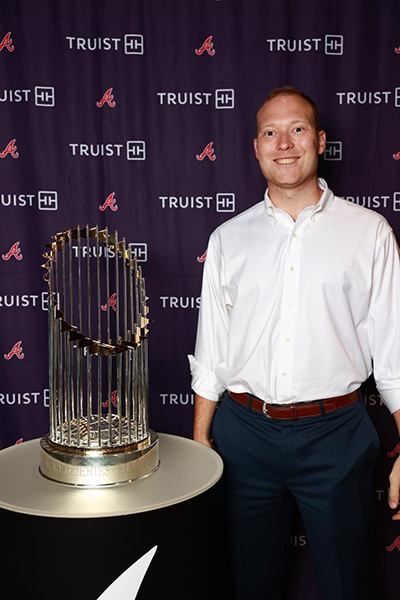 Man in a white shirt and dark pants smiles beside a tall silver trophy on a round table, with a purple TRUIST backdrop behind him.