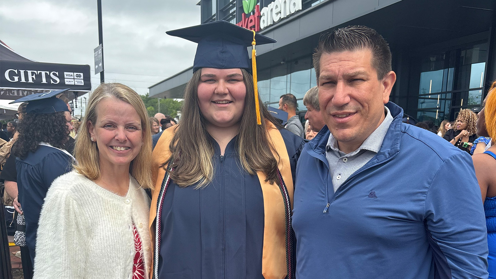 Graduate in navy cap and gown with gold tassel posing with two smiling adults outdoors at a graduation ceremony outside a building with a gifts tent nearby.
