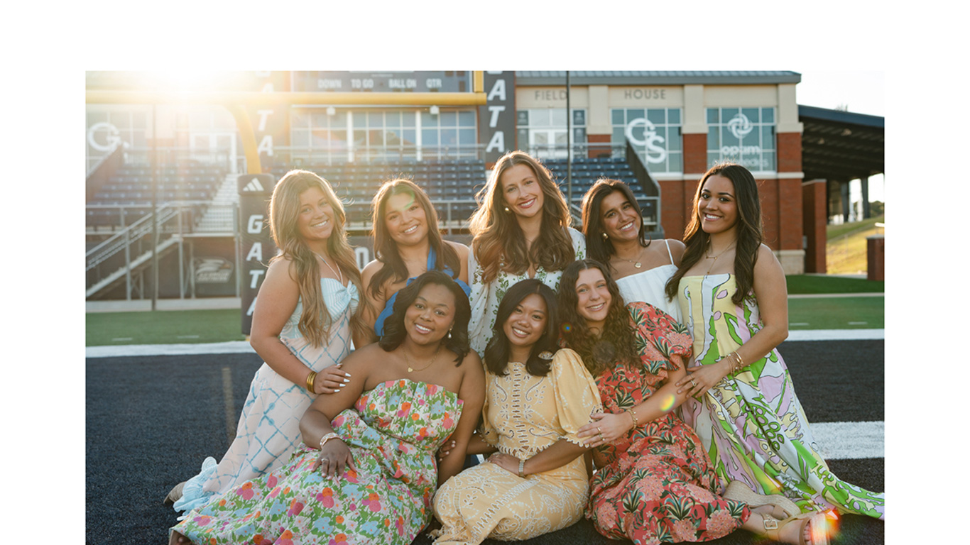 Group of eight young women in colorful dresses posing together on a football field, sunny stadium backdrop behind them.