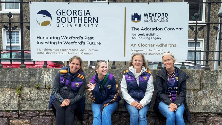 Four women wearing purple jackets sit on a stone ledge in front of banners for Georgia Southern University and Wexford European Campus.