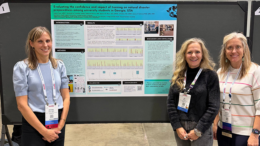 Three women researchers pose beside a large academic poster about training on natural disaster preparedness among university students in Georgia, USA, at a conference.