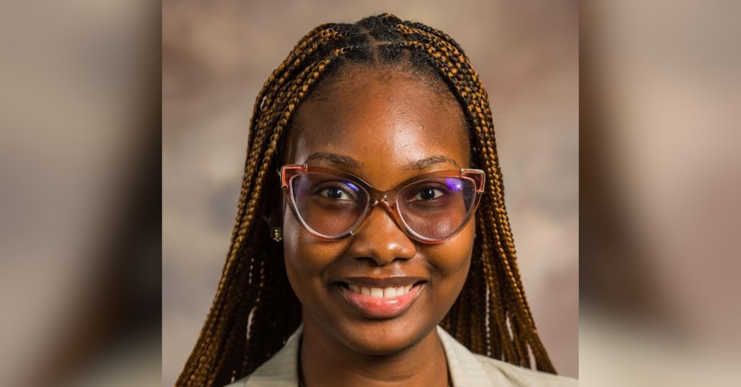 Smiling young woman with braided hair and pink-accented glasses wearing a striped beige blazer and a gold heart necklace against a neutral background.