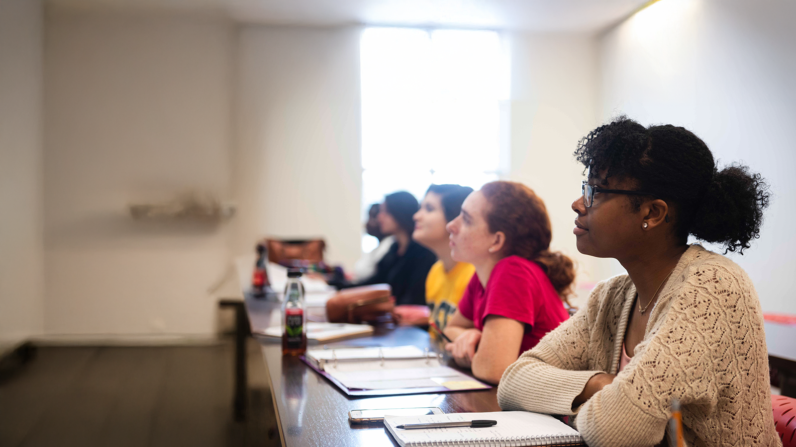 Group of students seated at a long desk listening to a presentation; foreground shows a woman in a beige knitted cardigan wearing glasses.