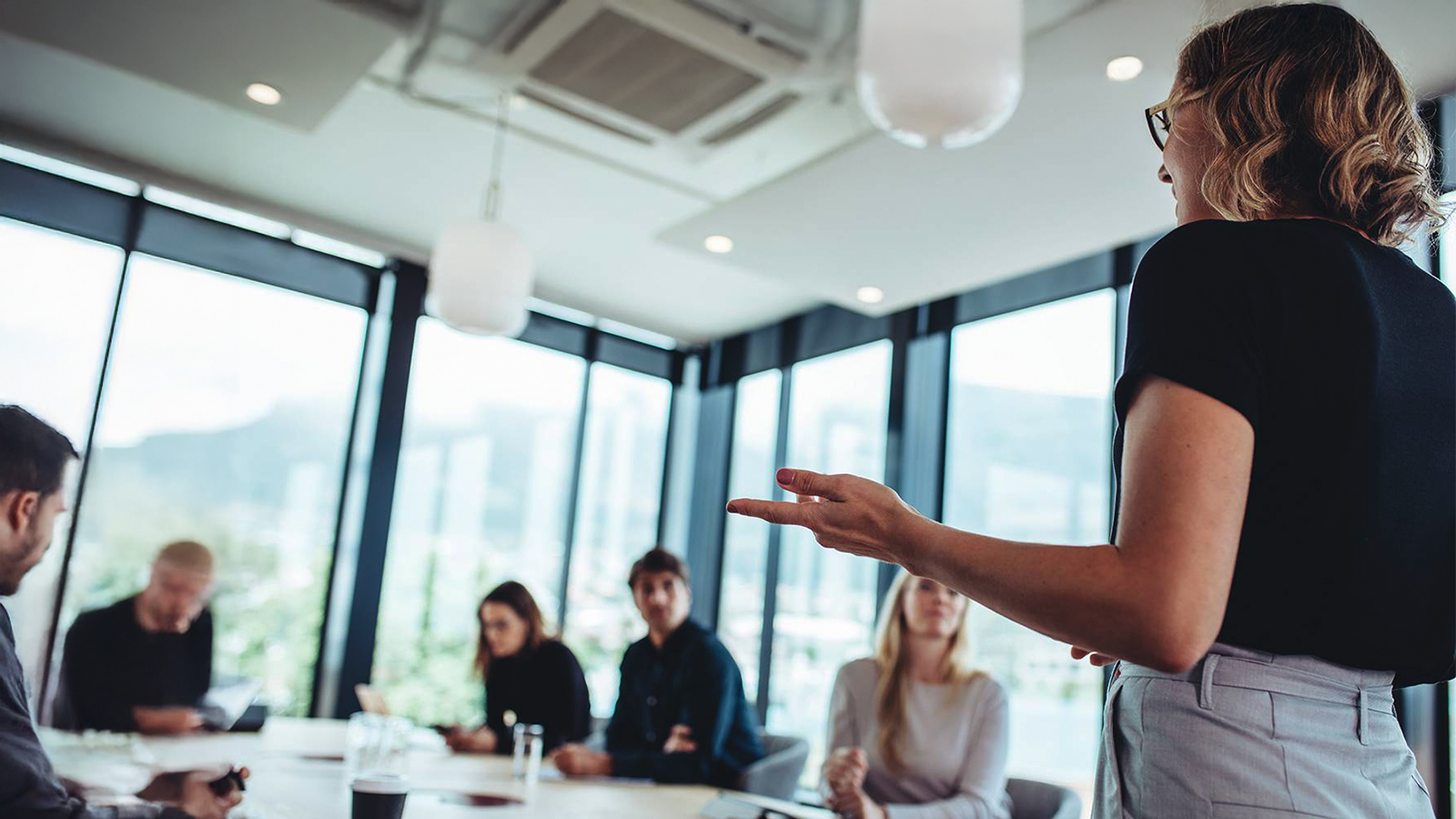 Woman presenting to a diverse group in a bright conference room, gesturing as others listen around a table.