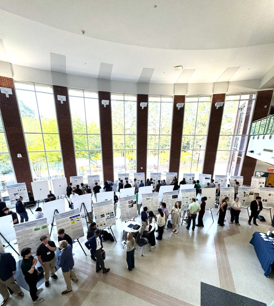 Students in the College of Engineering atrium standing beside research posters. The atrium has large windows shpwing trees.