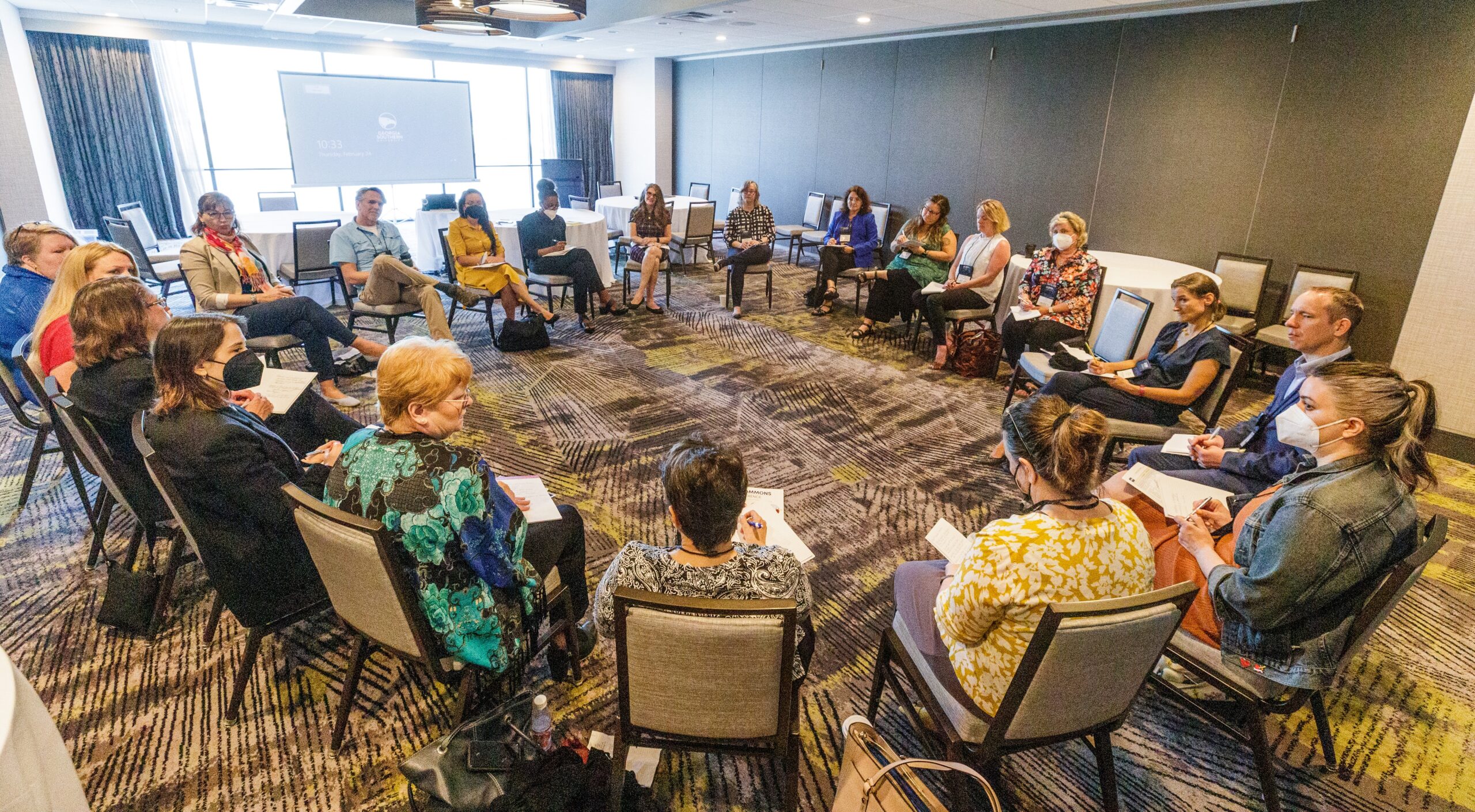 A circle of professionals in a conference room, some with notebooks, facing each other around a shallow U of chairs, projector screen in back.