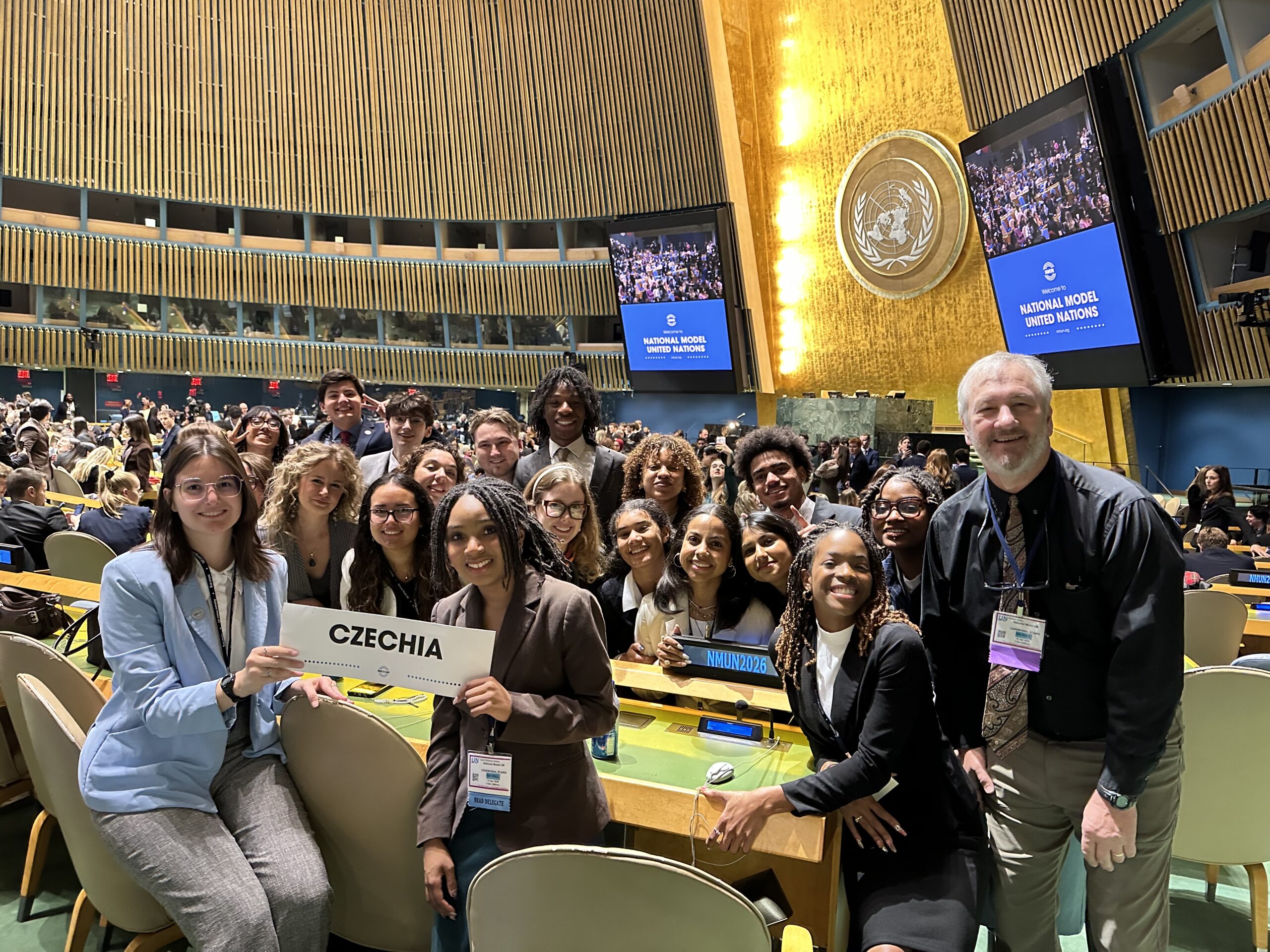 Diverse group of students taking a selfie in a UN conference hall, holding a Czechia sign.
