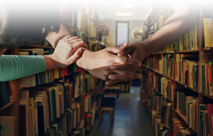 Three people join hands in the center of a library aisle, symbolizing teamwork and collaboration.