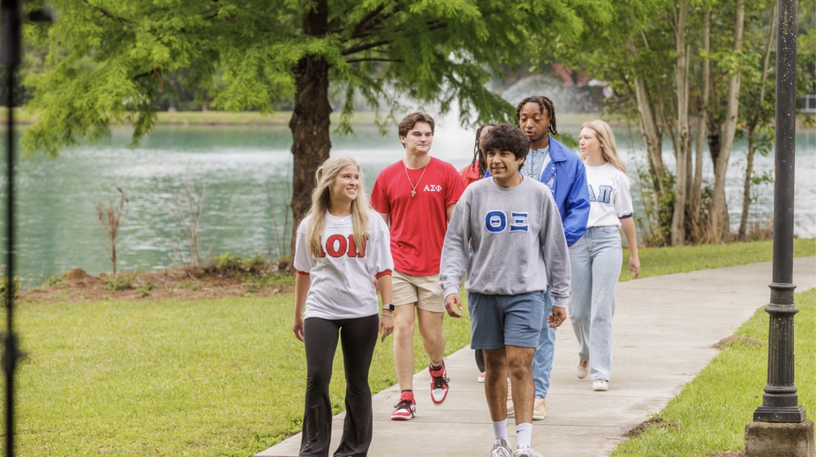 Group of five diverse young people walking together on a park path beside a lake, smiling.
