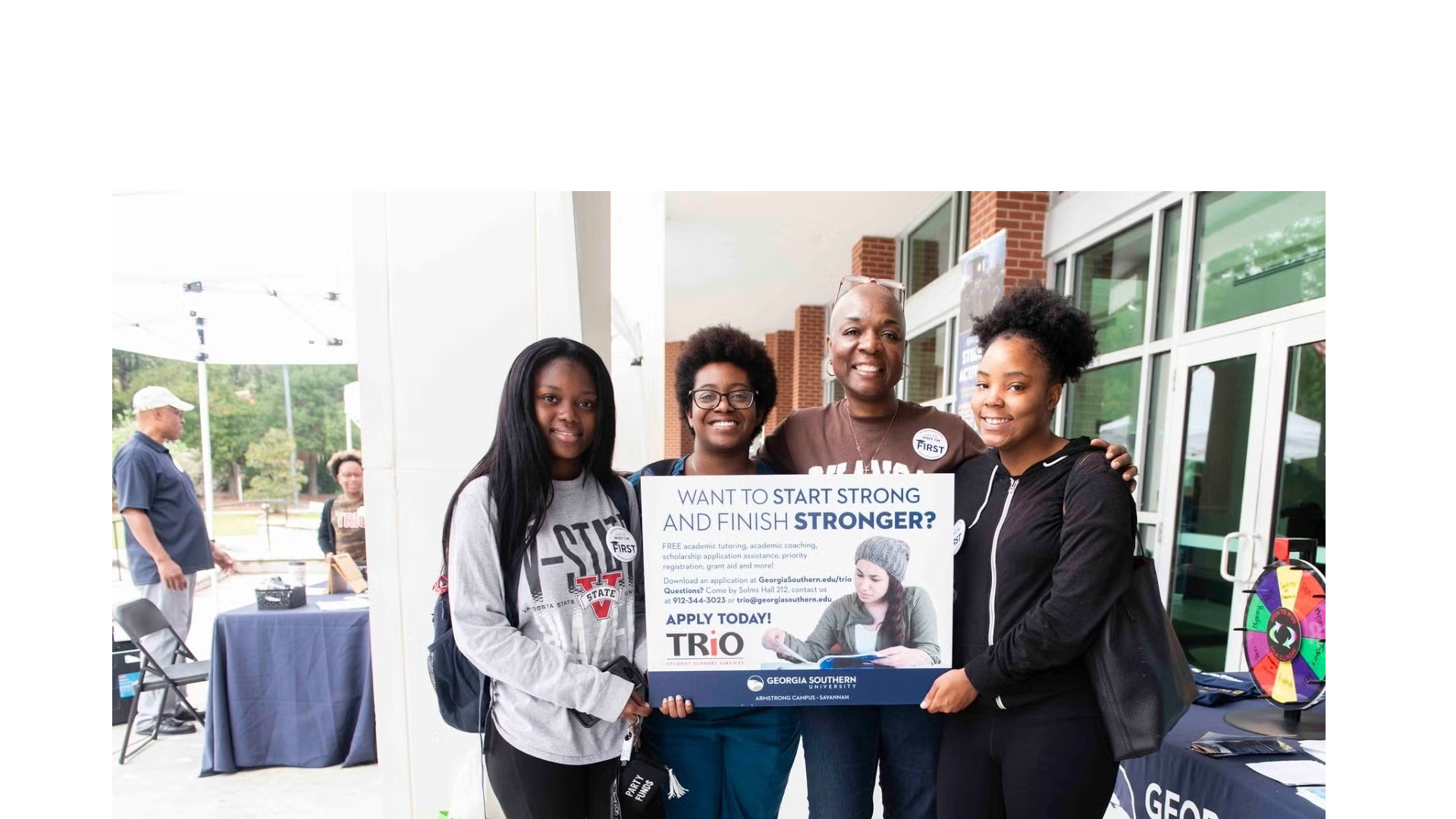 Four students smiling and posing with a TRIO program poster outside a building outside a campus fair setting?