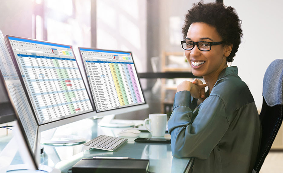 Woman with glasses sits at a desk with three monitors displaying spreadsheets, smiling at the camera.