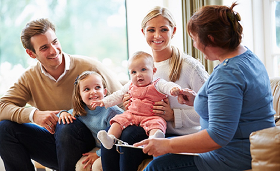 Family of four sitting on a sofa with a baby, smiling as a caregiver explains something with a clipboard in a bright living room.