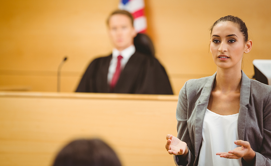 Young female attorney in a gray blazer speaks to the courtroom, with a judge seated behind.