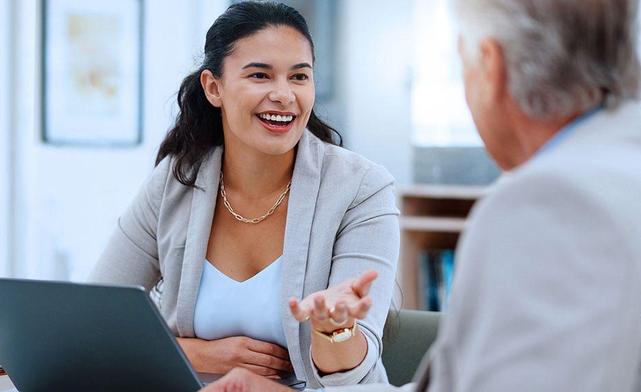 Professional woman smiling and gesturing while talking to an older man across a desk in a meeting room, laptop in view