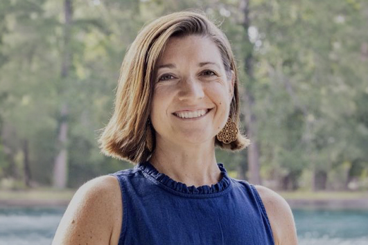 Woman with shoulder-length light brown hair smiling outdoors near a lake, wearing a blue sleeveless top and gold earrings.
