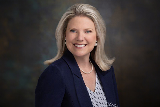 Professional headshot of a smiling blonde woman in a navy blazer and white blouse with a pearl necklace, against a dark studio backdrop.