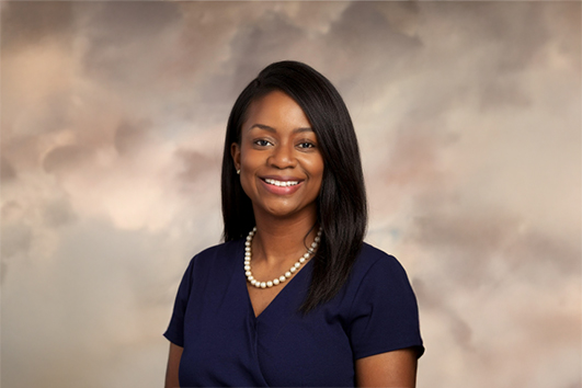 Professional headshot of a smiling Black woman in a navy blouse with a pearl necklace, against a soft neutral backdrop.