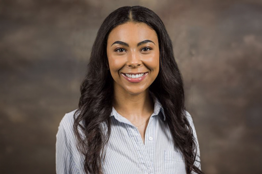Portrait of a smiling woman with long dark hair in a light blue striped shirt against a neutral studio backdrop.