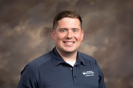 Smiling man in a navy polo shirt with a logo on the left chest, posed against a brown studio backdrop.
