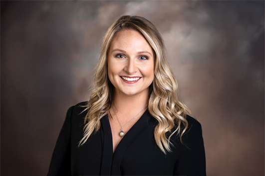 Professional headshot of a smiling blonde woman in a black blazer against a neutral studio background.