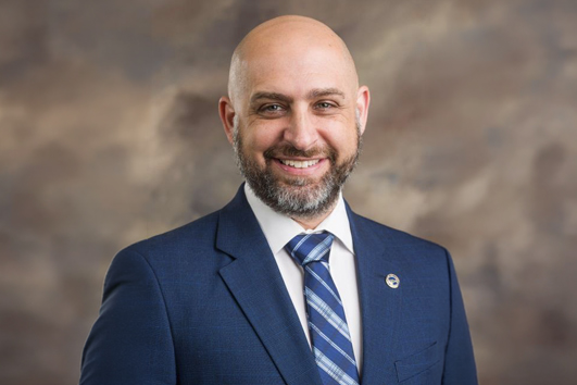 Professional headshot of a smiling bald man in a blue suit and striped tie against a neutral brown backdrop, suitable for a corporate bio.