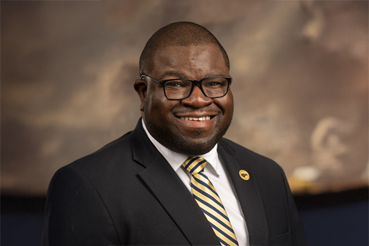 Smiling African American man in a dark suit with striped tie and glasses, posing for a professional headshot.