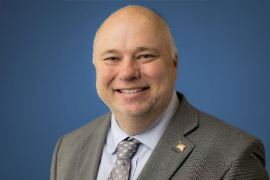 Smiling man in a gray suit and patterned tie against a blue background, with an American flag pin on his lapel.