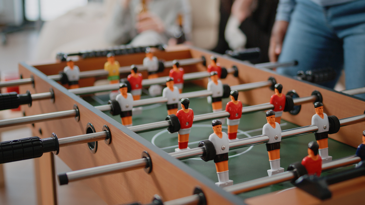 Foosball table with red and white figurines on metal rods, players in a game with blurred people in the background.