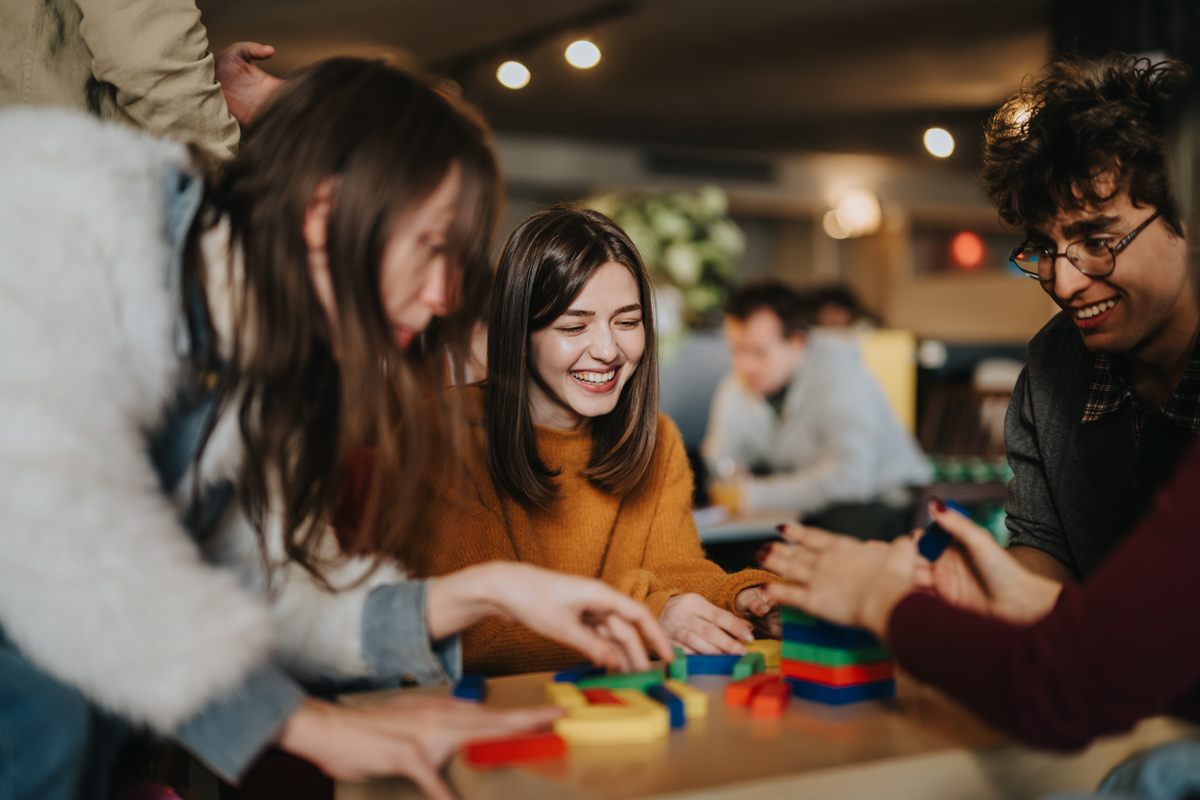 Group of young adults playing with colorful building blocks at a table, smiling and chatting.