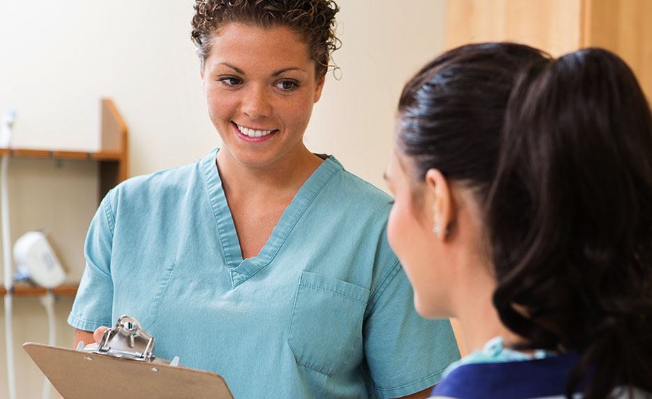Nurse in blue scrubs holds a clipboard and smiles at a patient in a clinic examination room.