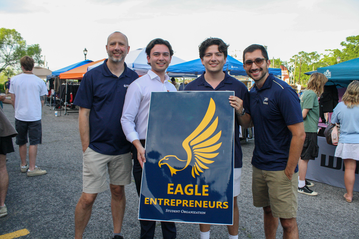 Four men at an outdoor fair pose with a large blue sign reading Eagle Entrepreneurs and a gold eagle logo.
