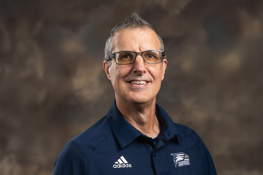 Smiling man with glasses in a navy polo shirt featuring Adidas and Georgia Southern logos, studio portrait