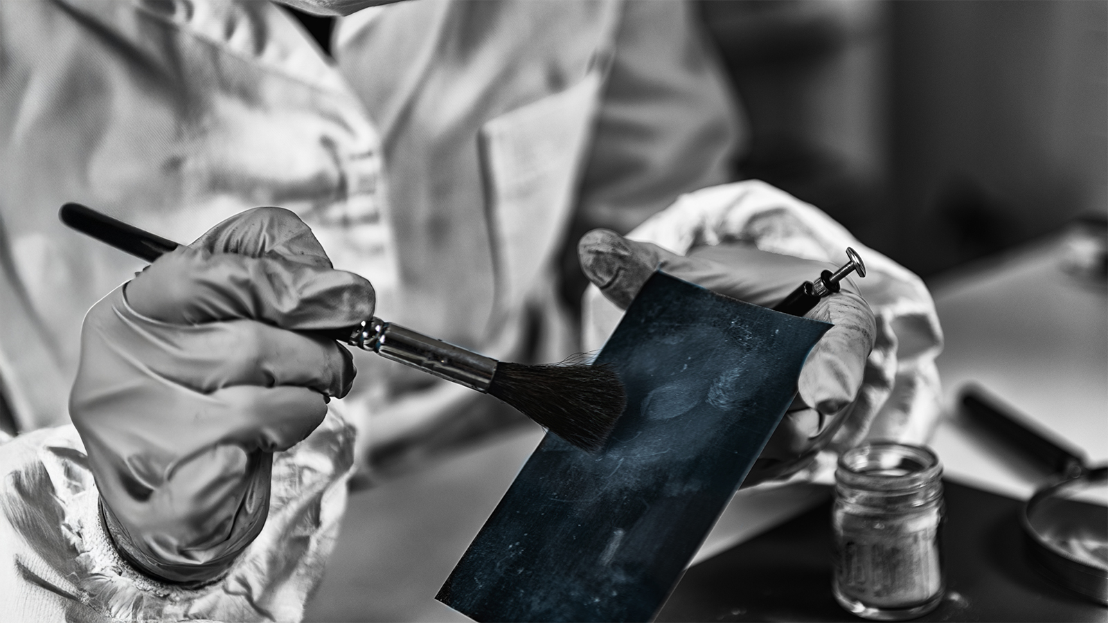 Person wearing gloves is painting a dark metal sheet with a brush in a workshop, with a jar of paint nearby