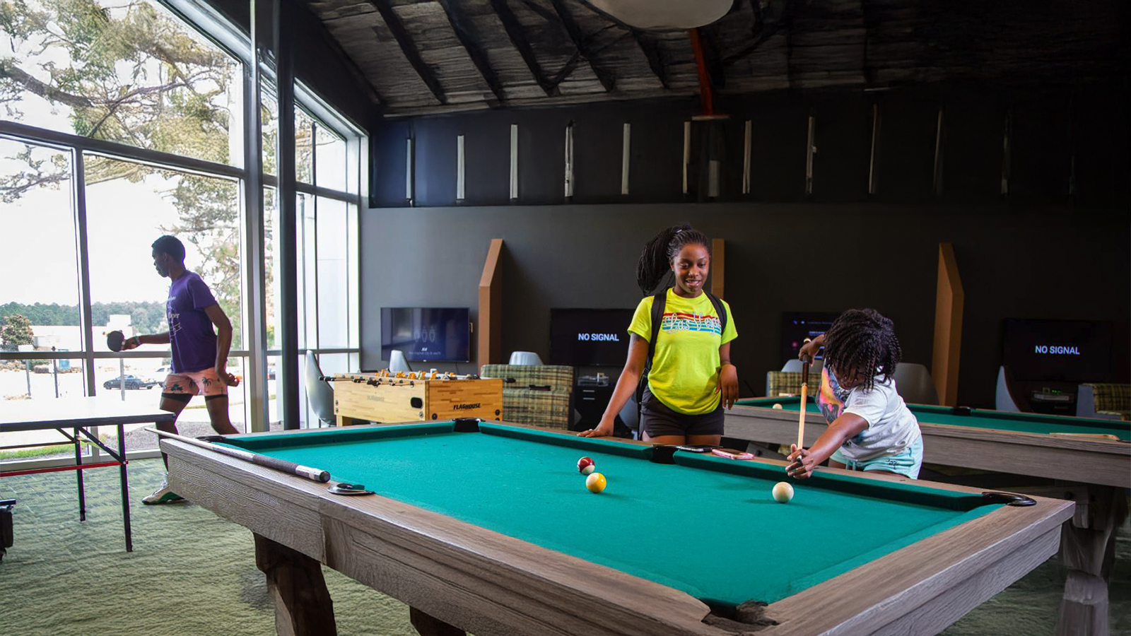 Children playing pool in a bright recreation room with large windows and a foosball table in the background, relaxed and social.
