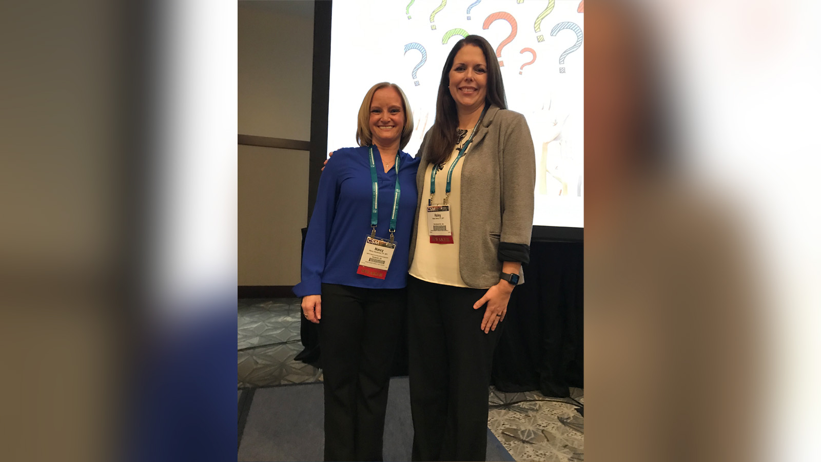 Two smiling women posing together at a conference, each wearing a lanyard with badge in front of a screen with colorful questions marks behind them.