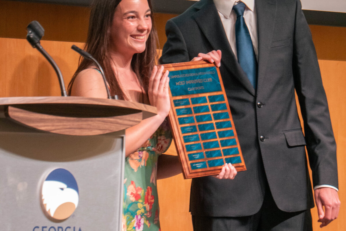 Smiling woman in a floral dress and a man in a suit pose on stage, holding a blue award plaque together on a Georgia Southern University podium scene.