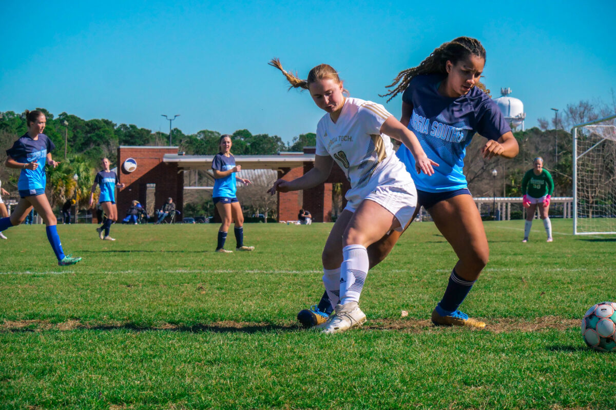 Girls' soccer match on a sunny day as players chase the ball near the goalpost on a grassy field.
