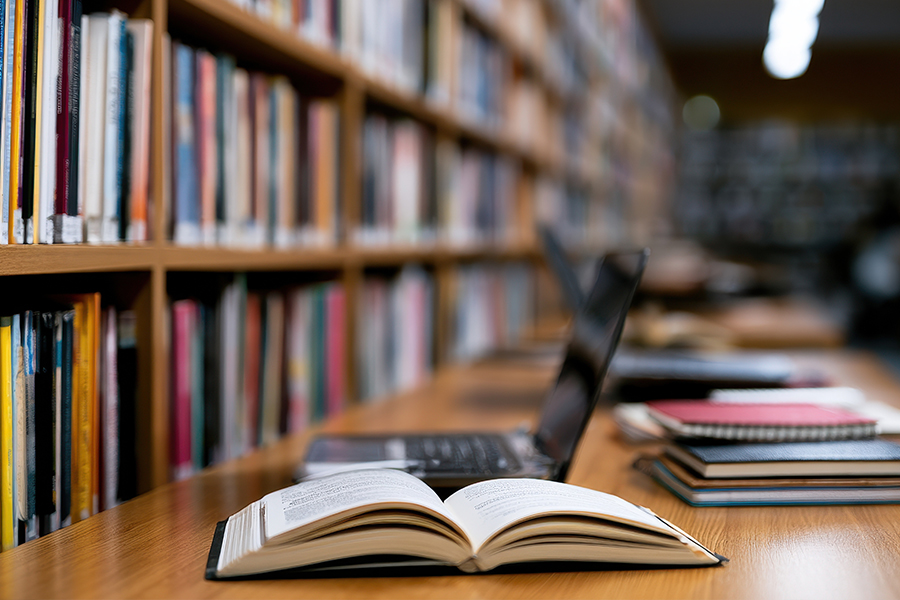 Open book spread on a wooden desk in a library, with a laptop and notebooks nearby.