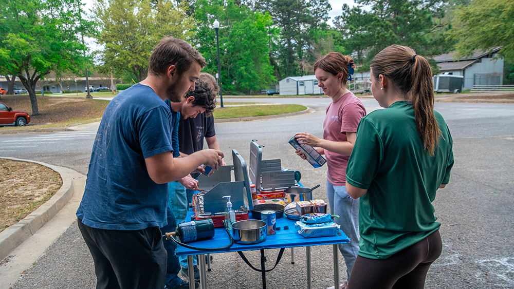 Five people gather around a blue folding table with cooking gear for an outdoor barbecue in a parking lot.