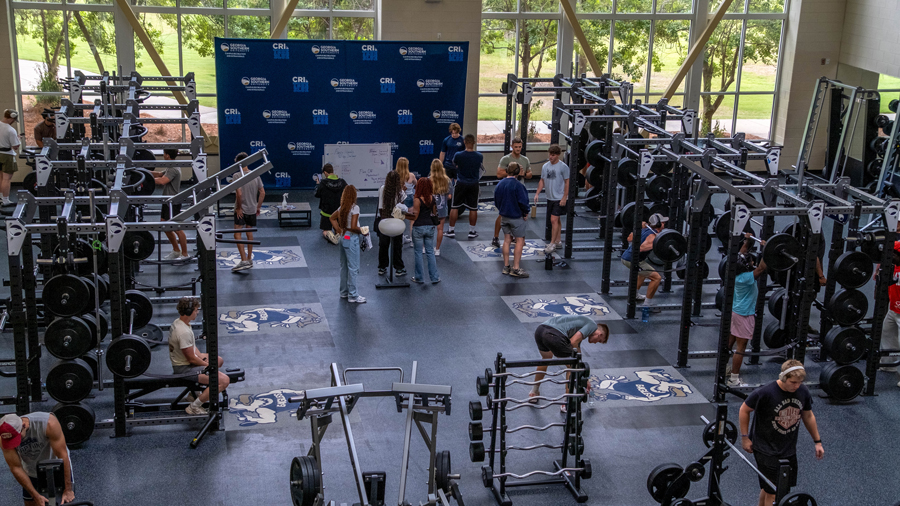 Gym with racks of barbells and weight plates; group of people gathered near a blue backdrop stage at the far end, likely a workshop or presentation.