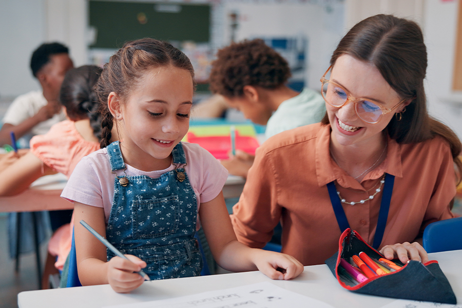 Girl in denim overalls drawing with a pencil at a desk, smiling teacher beside her in a bright classroom.