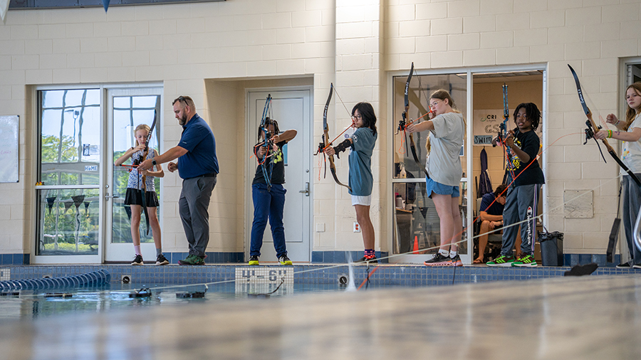 Instructor coaching children in archery along a pool deck inside a recreation center.