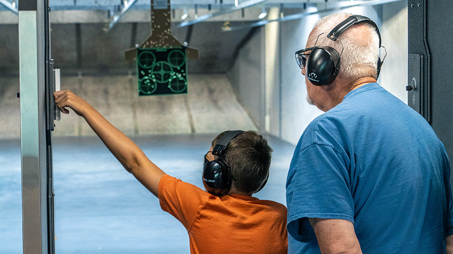 Boy in an orange shirt and an older man in a blue shirt at an indoor shooting range, both wearing hearing protection, as the boy reaches to operate a panel near a hanging target.
