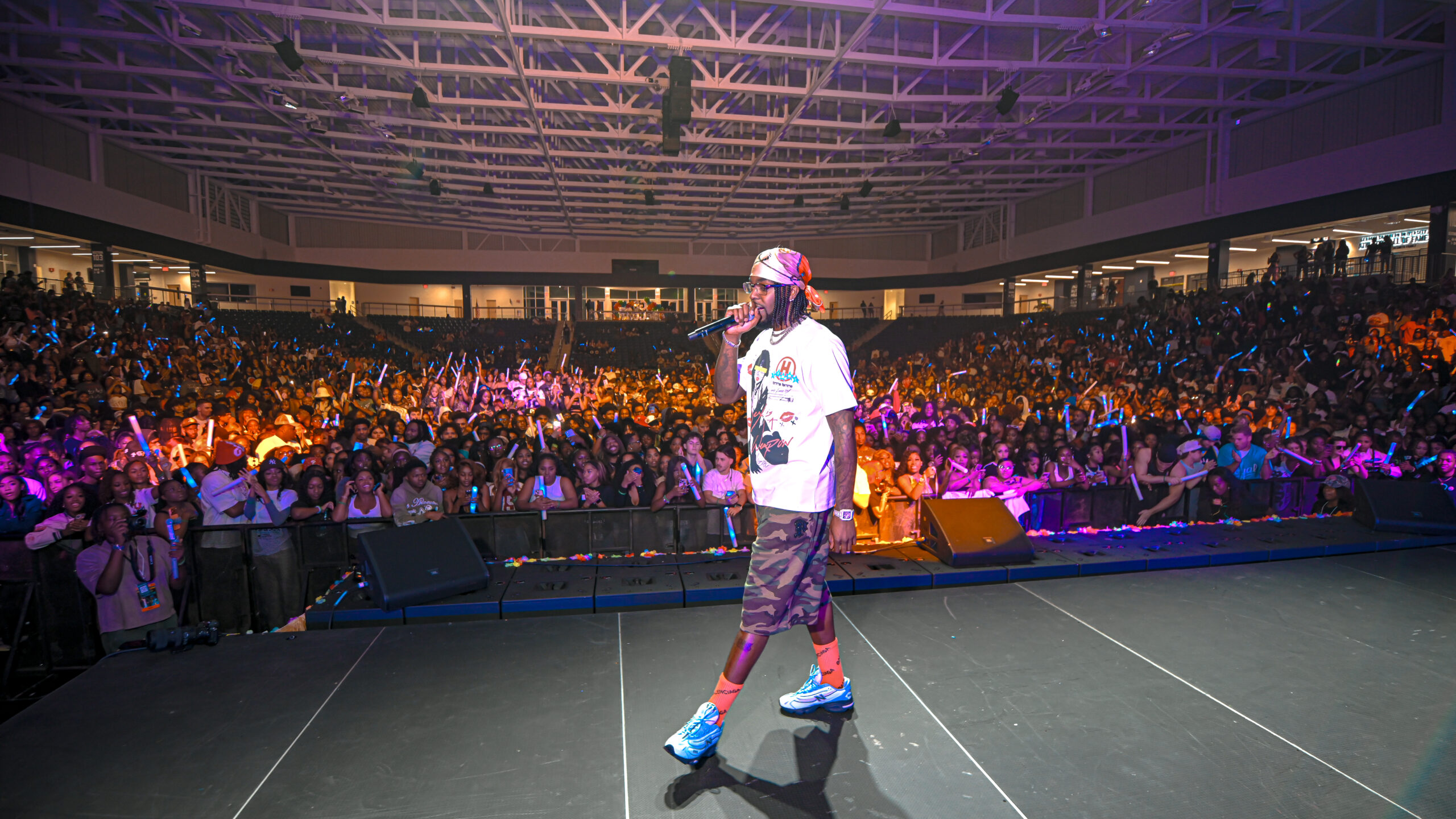 Live performer wearing white shirt and camo shorts sings into a microphone on a crowded arena stage, surrounded by fans with blue glow sticks.