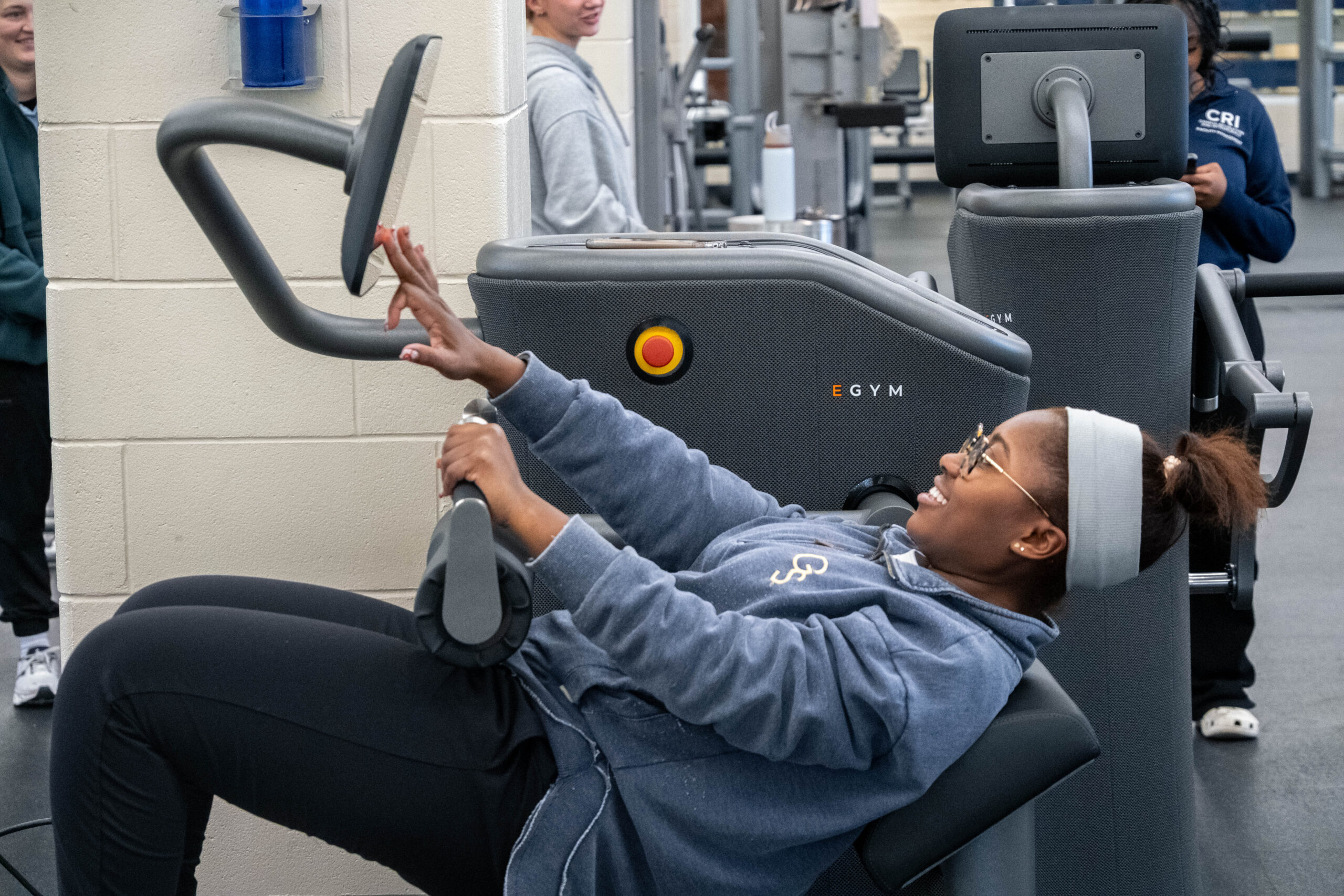 Woman in a gray hoodie reclining in a large adaptive gym chair, pushing the exercise handles in a fitness facility.
