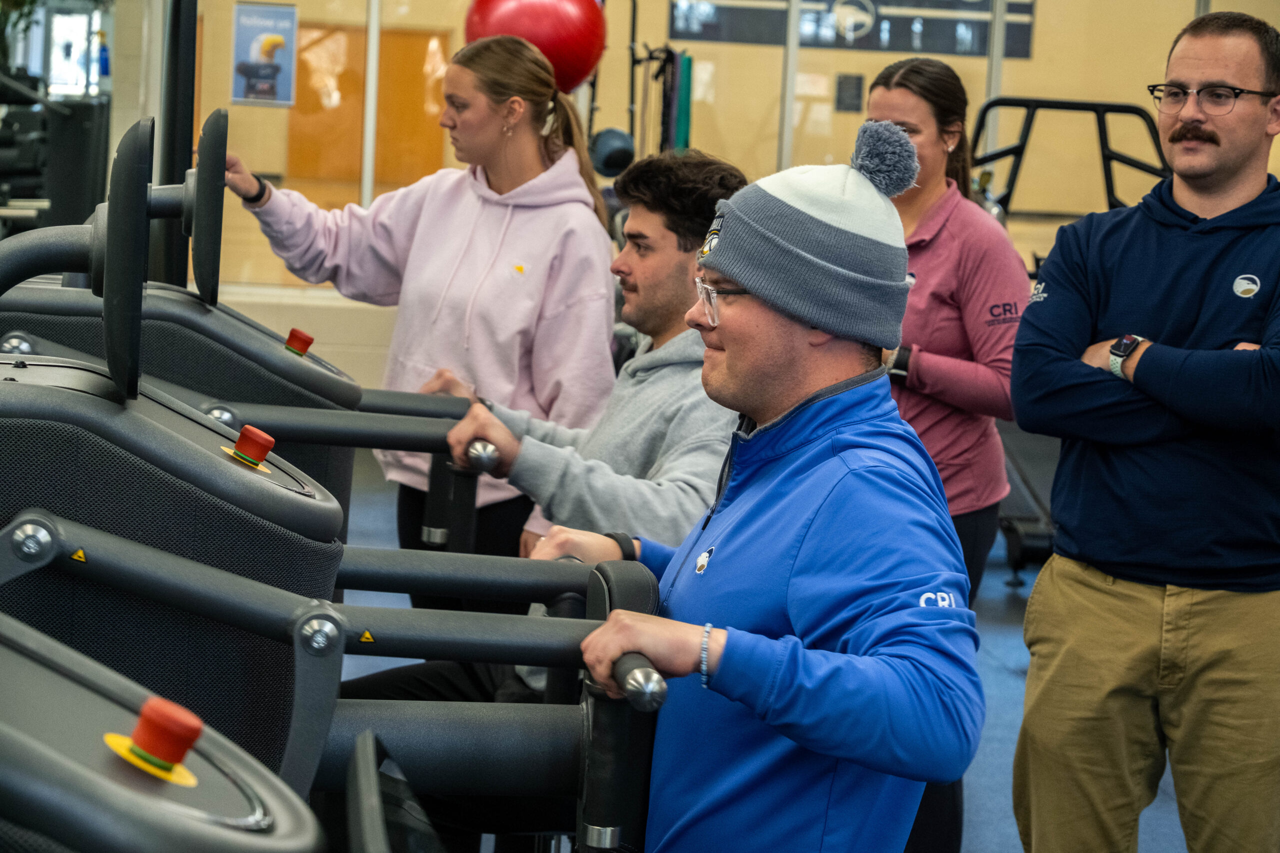 Group of people in a gym gathered around treadmills as a man in a blue jacket uses one treadmill in the foreground.