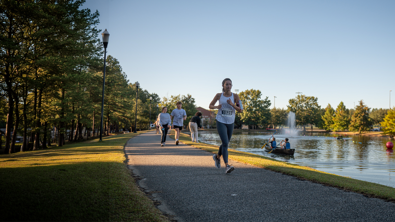 Female runner with bib 1523 jogging along a lakeside park path; others walk in the background as rowboats glide on the water and a fountain shoots up in the lake.
