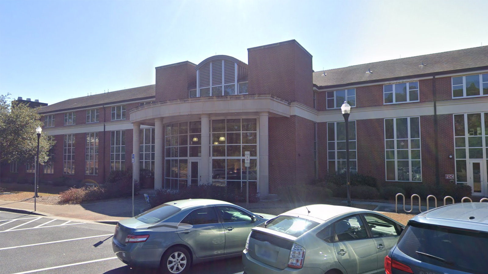 Brick institutional building with a large glass entrance and curved front, cars parked in the foreground on a sunny day.