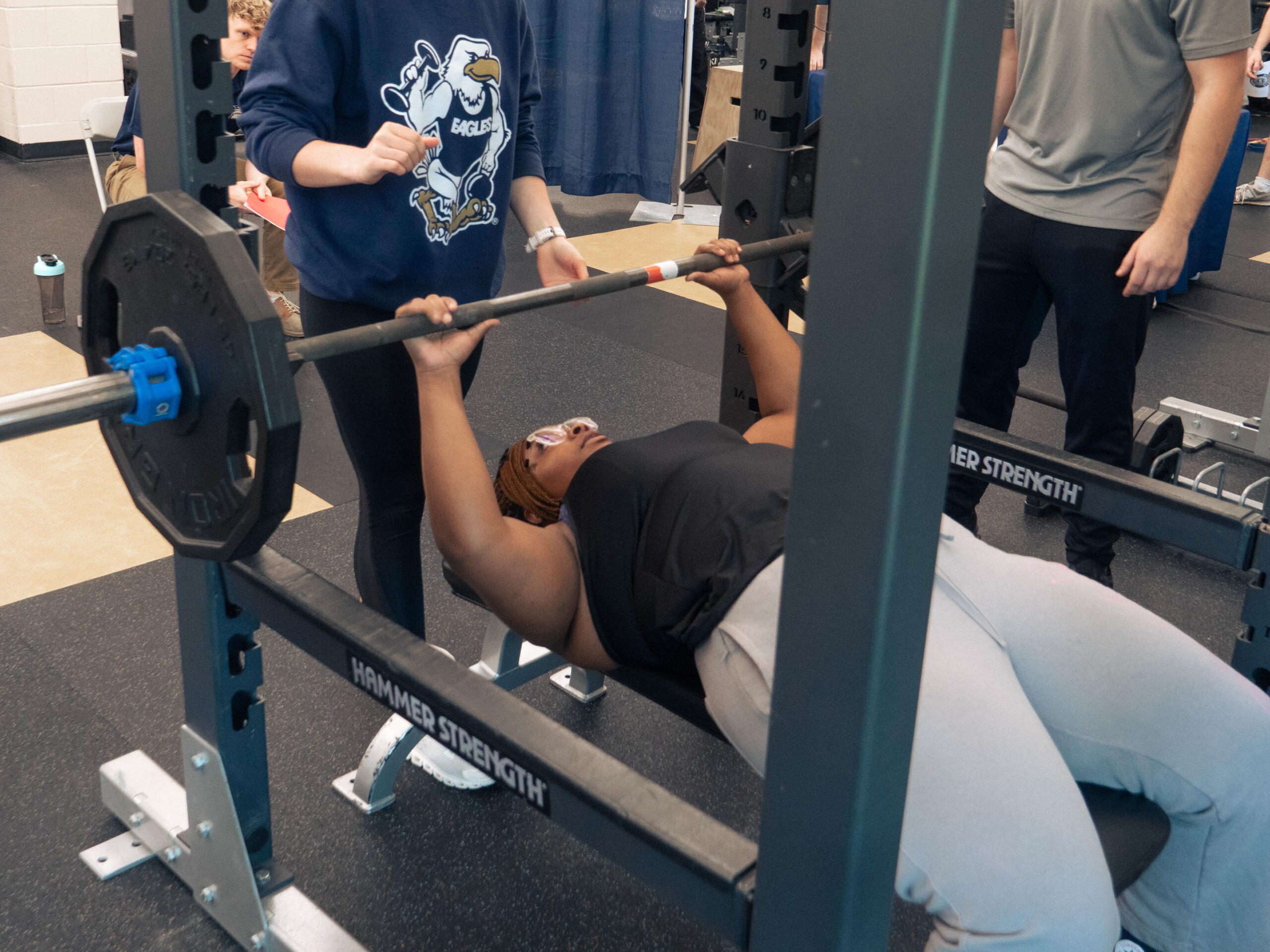 Person lying on a bench press, lifting a barbell while a spotter stands behind them in a gym setting (weight room)
