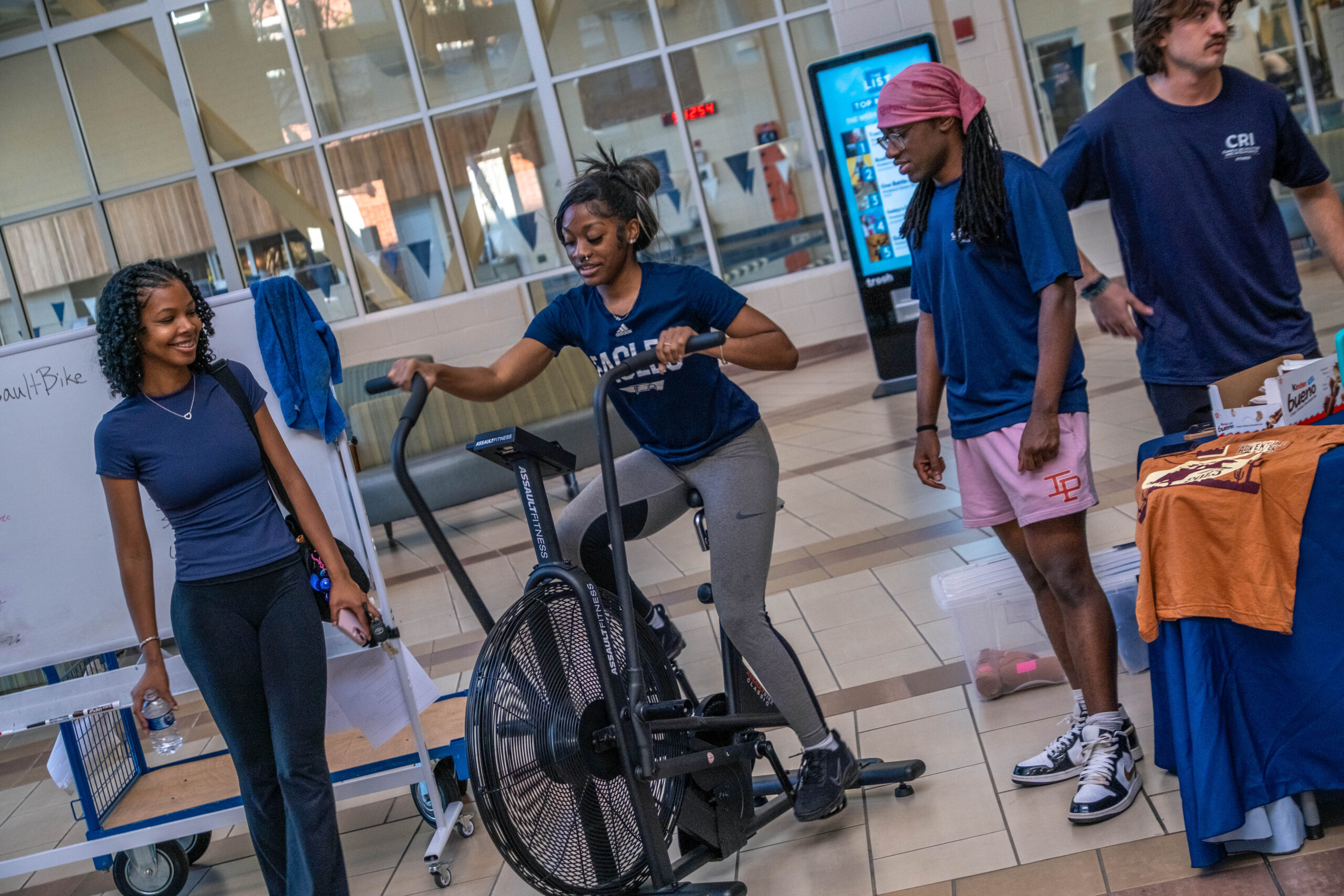 Young woman on a stationary bike demonstrates pedaling while others in blue shirts watch in a campus gym area.